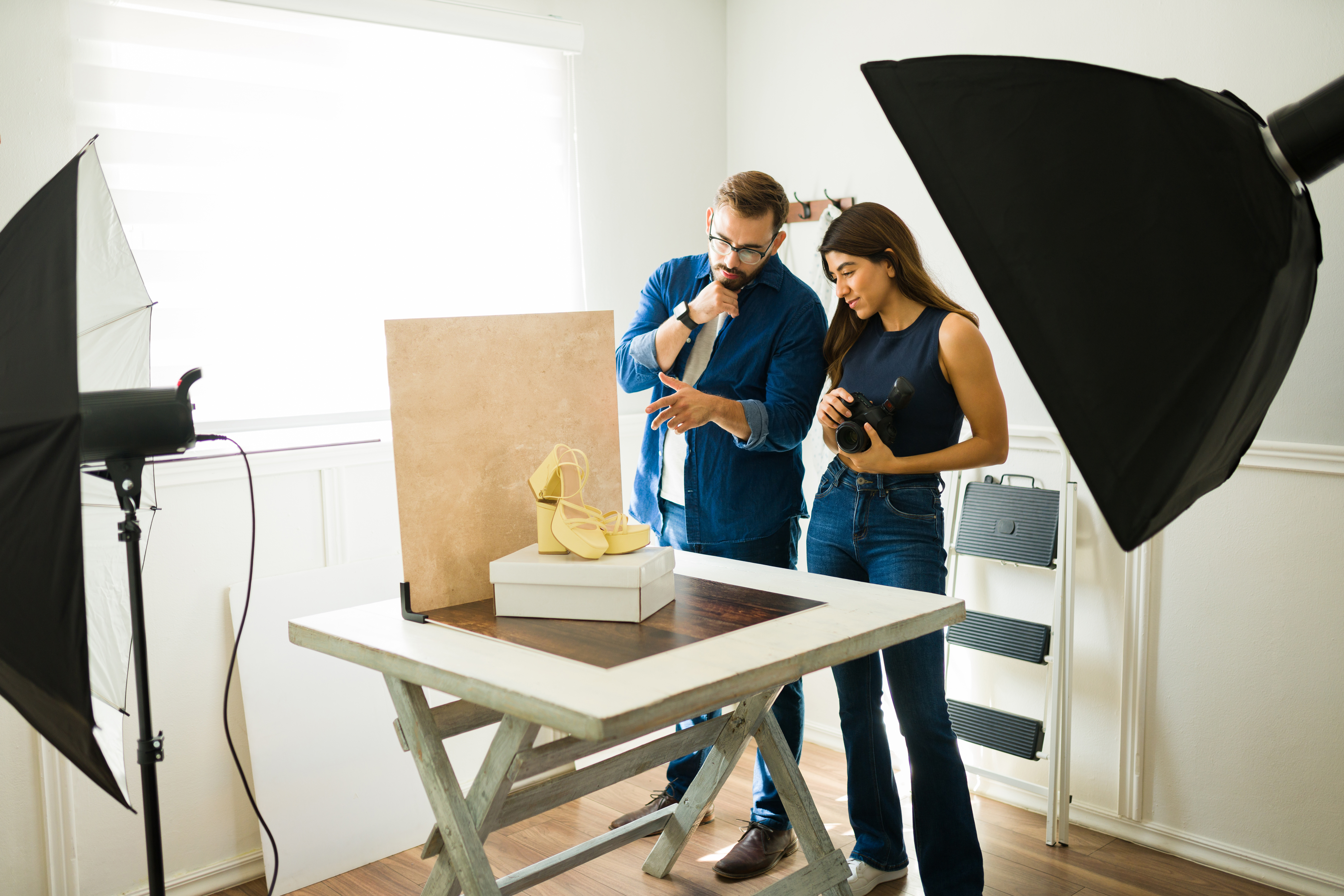 Two people in a photography studio setting examine a shoe displayed on a table. Photography equipment and lights are positioned around them.