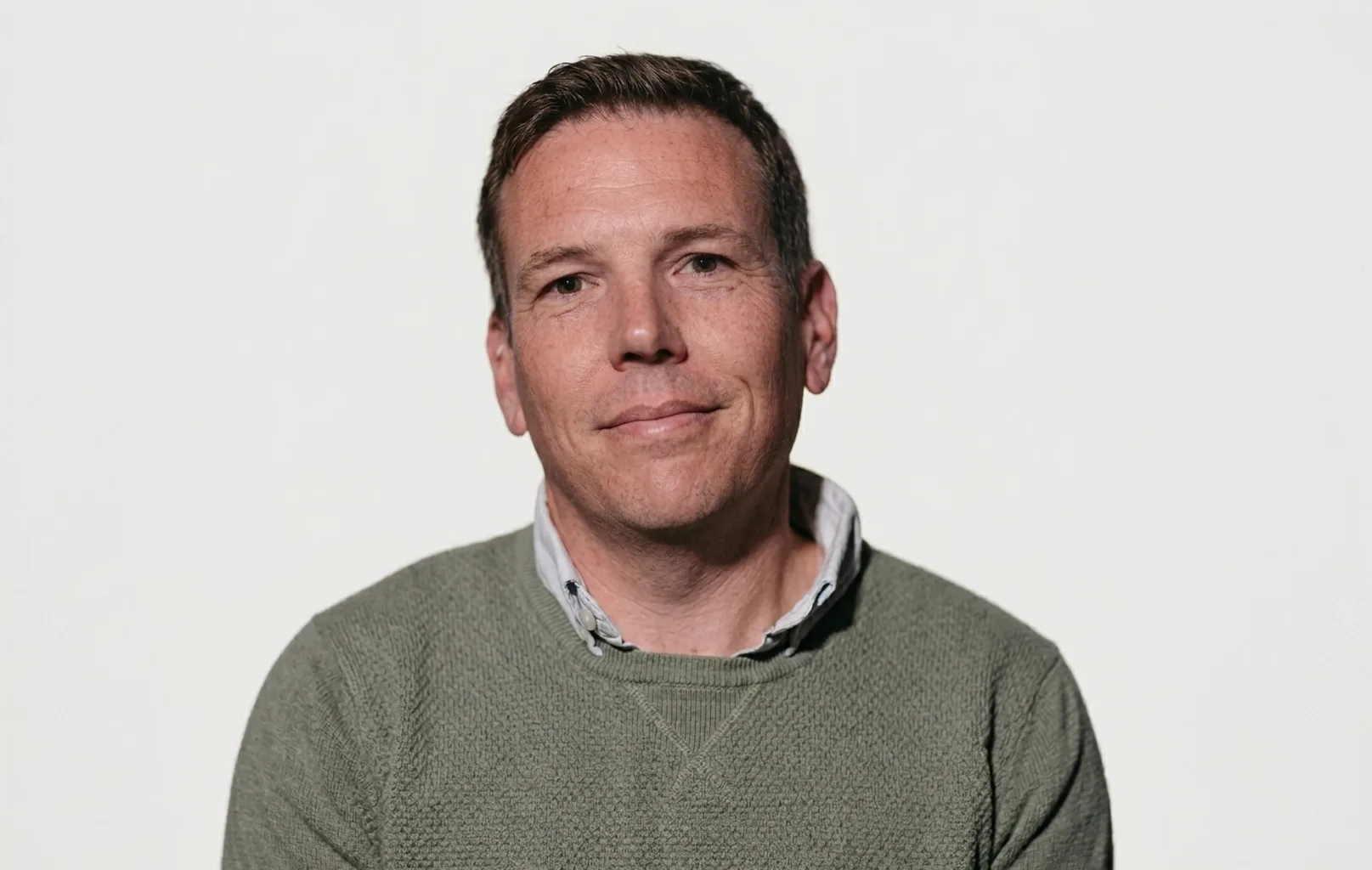A man with short brown hair, wearing a green sweater over a collared shirt, sits in front of a plain white background, looking at the camera.