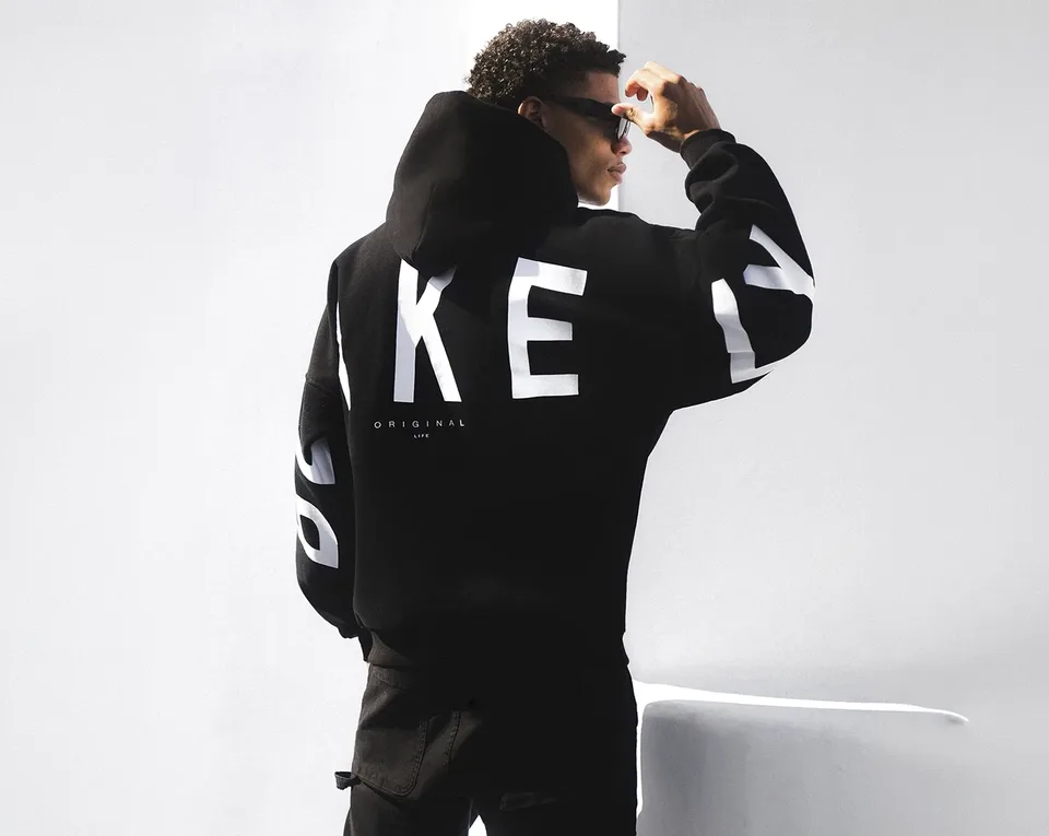 A person wearing a black hoodie with large white letters and black pants stands facing away, adjusting sunglasses in a minimal, well-lit room.