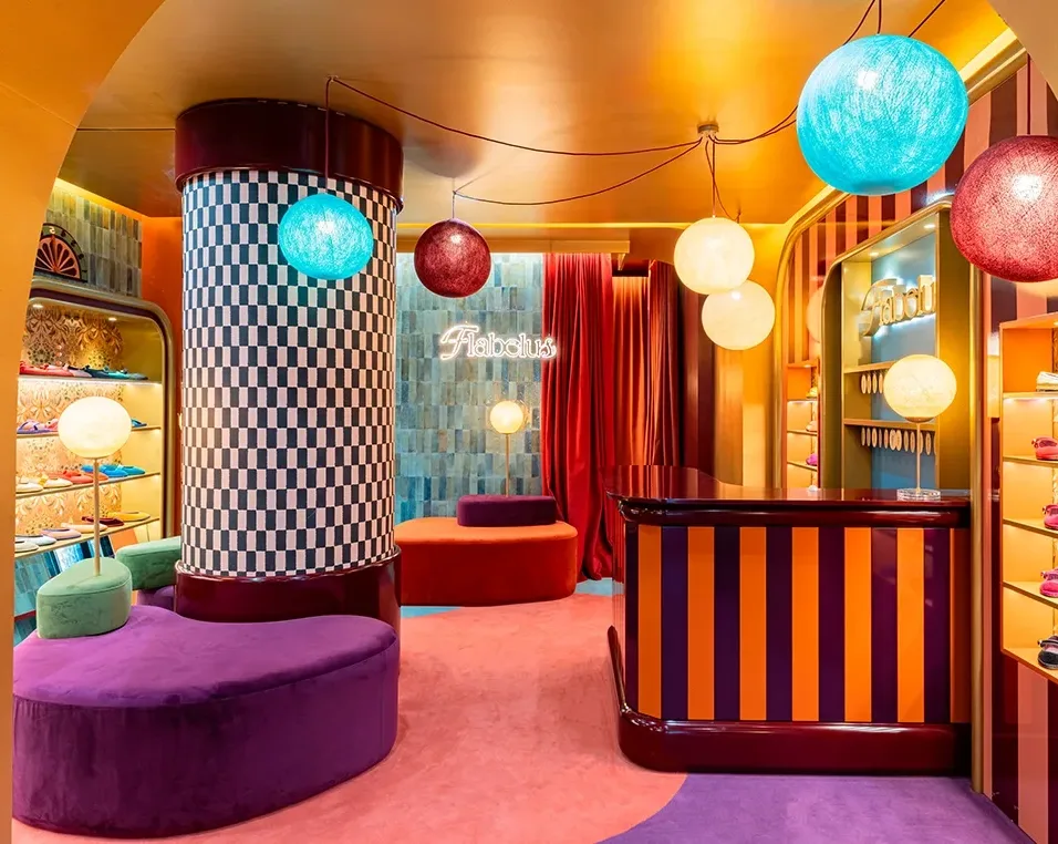 A brightly colored boutique interior with checkerboard and striped patterns, round hanging lamps, velvet furniture, and a neon "Fabulous" sign on the wall.