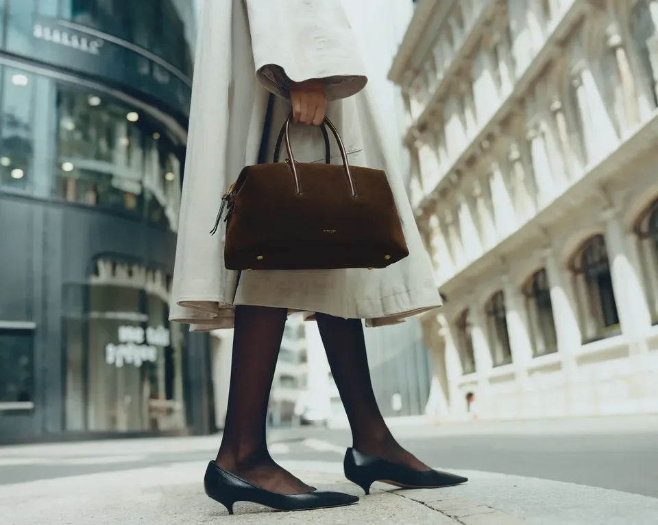 Person in a light-colored trench coat and black heels holding a dark brown handbag while standing on a city street.