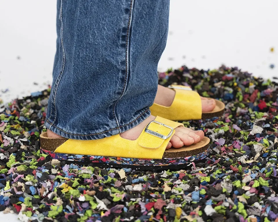 A person wearing yellow sandals and blue jeans stands on a pile of colorful shredded material scattered on a white surface.