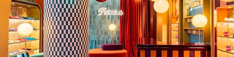 A brightly colored boutique interior with checkerboard and striped patterns, round hanging lamps, velvet furniture, and a neon "Fabulous" sign on the wall.