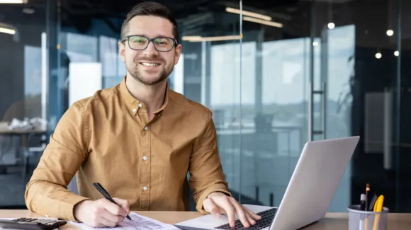Man with glasses and a brown shirt sits at a desk, smiling, holding a pen and working on a paper beside an open laptop in a modern office setting.