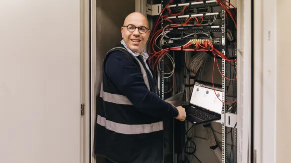 A person wearing glasses and a reflective vest works on a laptop connected to a server rack with multiple cables in a server room.