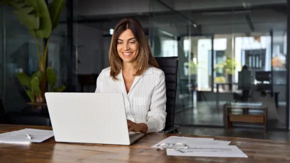Woman in business attire smiling while working on a laptop at a desk in a modern office, with documents and eyeglasses nearby.
