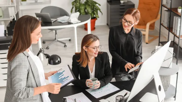 Three women collaborate in a modern office, reviewing charts and working at a computer. One holds a clipboard with graphs, another sits with a mug, and the third types on a keyboard.