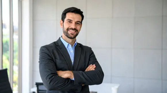 A man in a suit stands indoors with his arms crossed, smiling, in front of a light-colored wall and window.