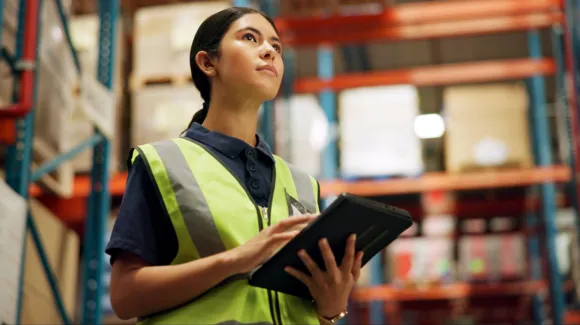 A woman in a reflective vest holds a tablet and looks upward in a warehouse with shelves and boxes in the background.