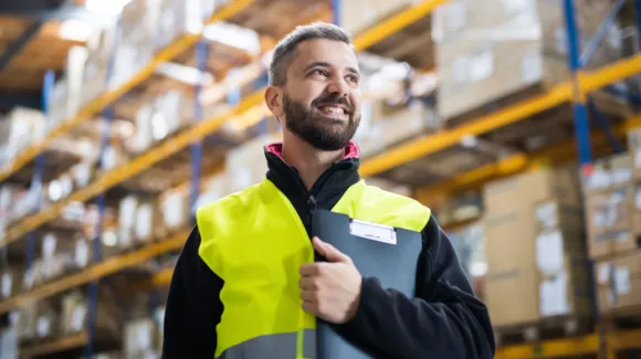 A man in a high-visibility vest holds a clipboard and stands in a warehouse with shelves of boxes in the background.
