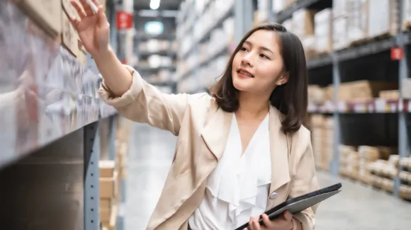 Woman standing in a warehouse aisle, reaching for a box on a shelf while holding a tablet in her other hand.