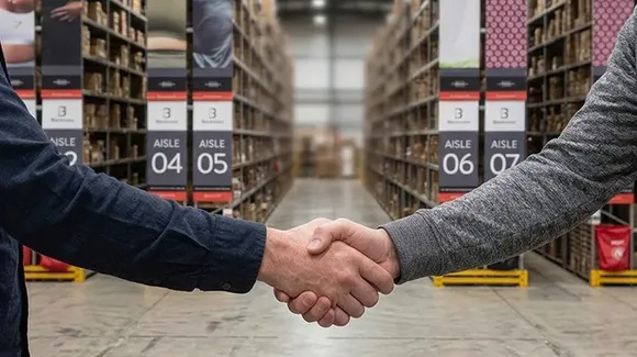 Two people shake hands in a warehouse, standing between aisles filled with stacked shelves.