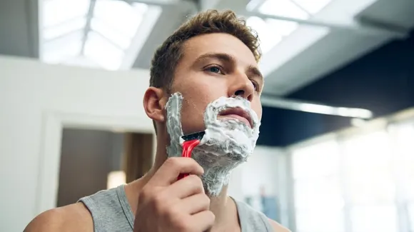 A man in a tank top shaves his face with a razor in a brightly lit bathroom. His face is covered in shaving cream.