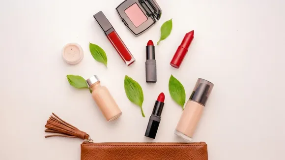 Assorted makeup items and green leaves arranged on a white surface with a brown makeup bag at the bottom.