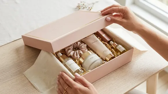 A person opens a pink gift box containing skincare products, a scrunchie, and shredded paper filler on a light wooden table near a window.