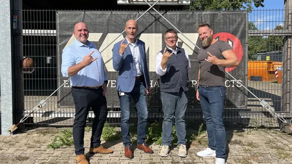 Four men standing in front of a branded fence, all making rock-and-roll hand gestures.