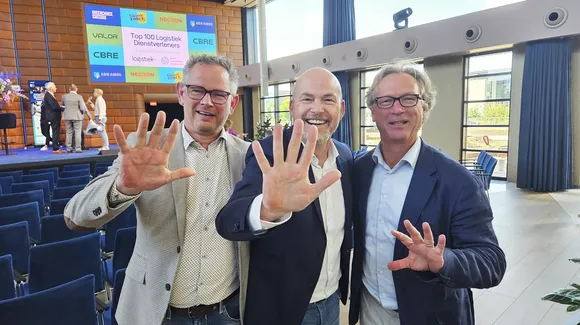 Three men in business attire stand together smiling and holding up five fingers each in a conference room with a stage and large screen in the background.