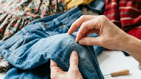 Hands repairing a tear in blue denim jeans with a needle and thread, with various clothing items in the background.