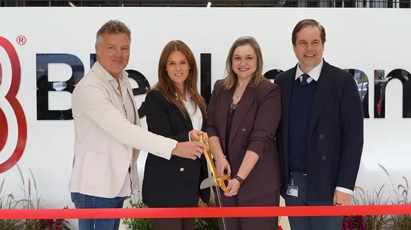Four people stand in front of a company sign, two women in the center holding large scissors, preparing to cut a red ribbon at an event.