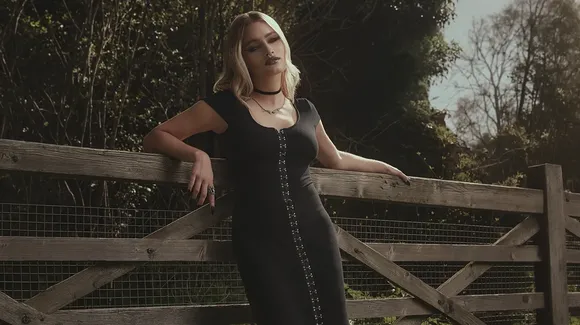A woman in a black dress with silver buttons stands outdoors, leaning against a wooden fence with trees and foliage in the background.