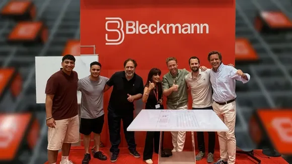 Seven people stand together smiling in front of a red Bleckmann display booth, with a white table and a blurred background featuring the company logo.