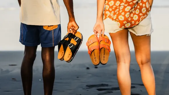 Two people stand side by side on a beach, holding sandals in their hands; one person wears blue shorts, the other wears white shorts and a floral top.