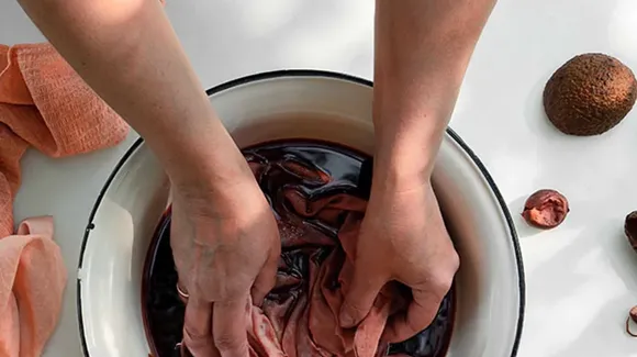 Two hands submerge pink fabric in a bowl of dark liquid, with pieces of avocado and a towel on a white surface nearby.