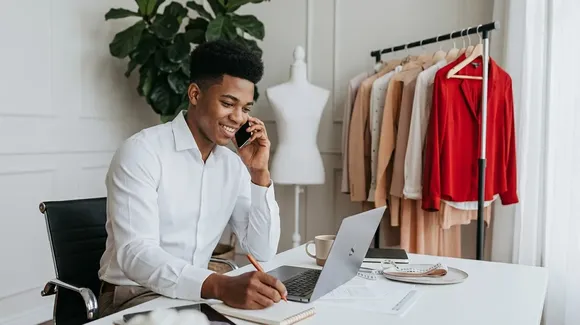 A man in a white shirt talks on the phone and writes in a notebook while working on a laptop at a desk. Clothes are hanging on a rack behind him.