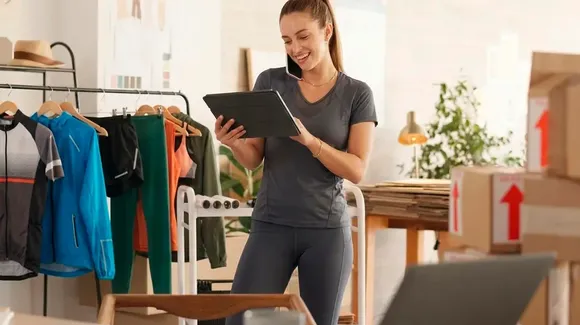 A woman in athletic wear smiles while holding a tablet and talking on a phone in an office with clothing racks and shipping boxes.