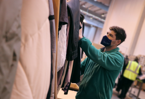 A person in a green jacket and mask organizes clothes on a rack in a warehouse setting.