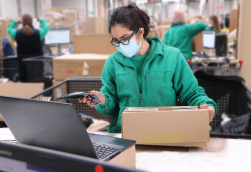 Person in a green fleece and mask scans a package next to a laptop in a warehouse setting. Others work in the background with computers and boxes.