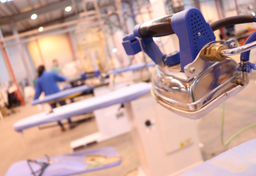 Industrial iron on ironing table in a factory setting, with blurred background showing workers and equipment.