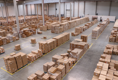 Rows of stacked cardboard boxes in a large, organized warehouse with high ceilings and concrete floors.