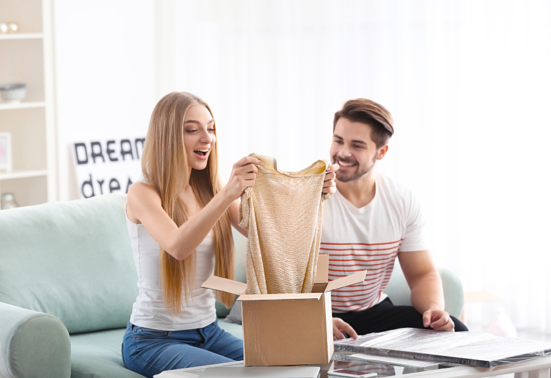 A man and woman seated on a sofa, unpacking a package with fashion