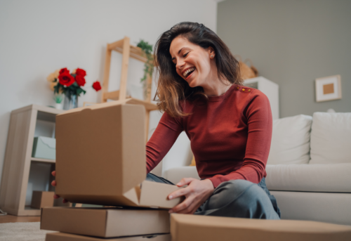 A woman sits on the floor in a living room, smiling as she opens a cardboard box with several other boxes stacked nearby.