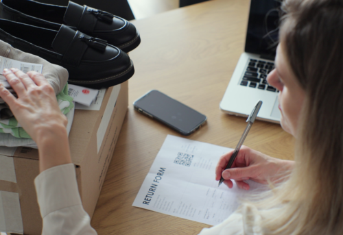 A woman fills out a return form next to a box with clothes and shoes, a smartphone, and a laptop on a wooden table.