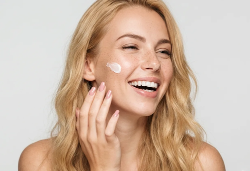 Smiling woman with long blonde hair applies a dab of cream to her cheek against a plain light background.