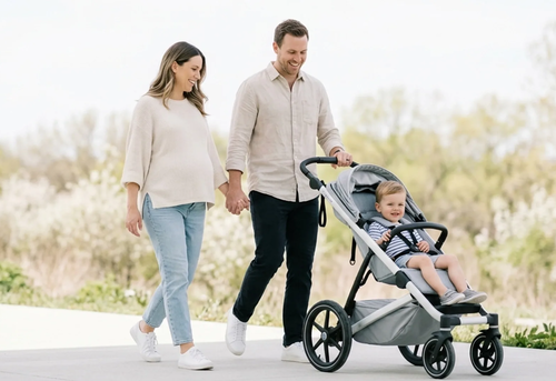 A man and woman walk outside holding hands while pushing a young child in a stroller on a paved path.