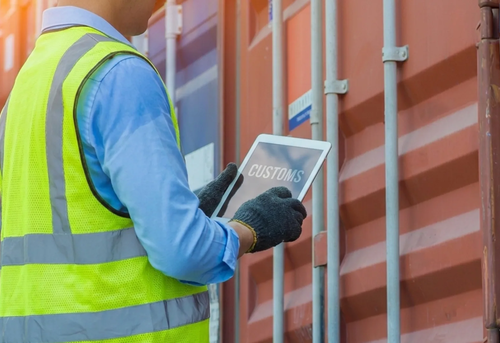 A person wearing a safety vest and gloves holds a tablet displaying "CUSTOMS" while inspecting a shipping container.