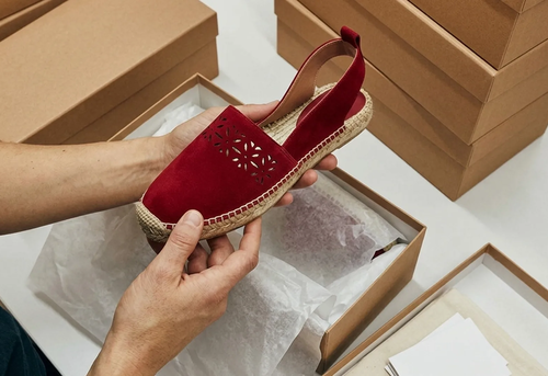 Hands holding a red espadrille with a cut-out pattern above an open shoe box, surrounded by other closed cardboard boxes.