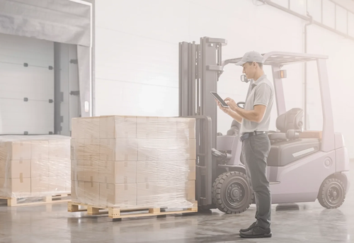 A worker stands beside a forklift and checks a clipboard near a large wrapped pallet inside a warehouse.