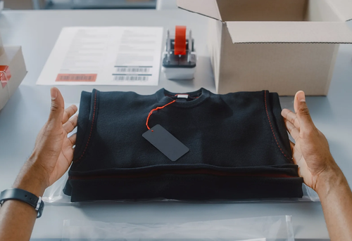 A person prepares to pack a folded black sweatshirt with a blank tag into a shipping box on a table with packing supplies and labels.