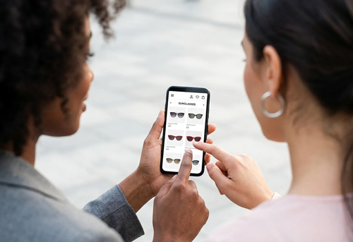 Two people look at a smartphone screen displaying a shopping website with various pairs of sunglasses, while one person points at a pair.