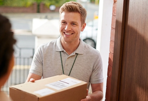 A delivery person wearing a light gray polo shirt hands a cardboard package to someone at a doorway, smiling.