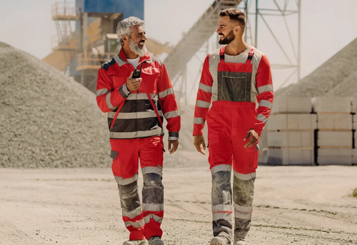 Two construction workers in red uniforms walk side by side at a gravel site, talking and smiling, with industrial equipment visible in the background.