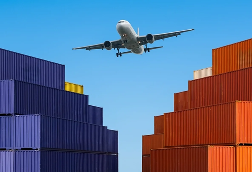 A commercial airplane flies over stacked blue and orange shipping containers against a clear blue sky.