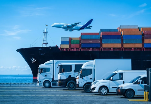 A large cargo ship with containers, several delivery trucks parked in front, and an airplane flying overhead against a blue sky.