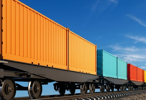 A freight train with orange, teal, blue, yellow, and red shipping containers travels on railroad tracks under a clear blue sky.
