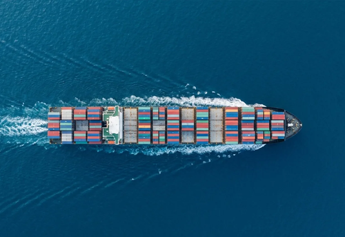 Aerial view of a cargo ship carrying stacked shipping containers moving through calm blue ocean water.