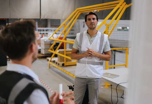 Two men in a warehouse, one wearing a safety vest and the other partially visible in the foreground.